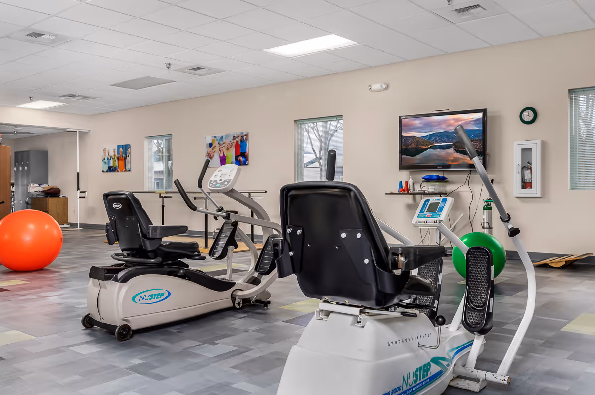 Interior view of a fitness room in a senior living facility with exercise equipment including two recumbent exercise bikes, a large orange exercise ball, and a green exercise ball. The room has light-colored walls, windows, a wall-mounted TV displaying a scenic landscape, and some fitness accessories on a shelf beneath the TV.