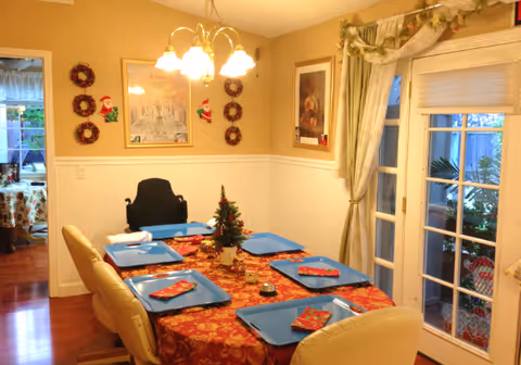 A dining room decorated for Christmas with a table set for six people. The table has a red and gold patterned tablecloth, blue trays, and Christmas napkins. A small decorated Christmas tree is in the center of the table. The room has cream-colored walls with white wainscoting, a chandelier with five lights hanging above the table, and a large window with curtains on the right side. There are Christmas wreaths and Santa decorations on the wall, and a doorway leading to another room is visible on the left.