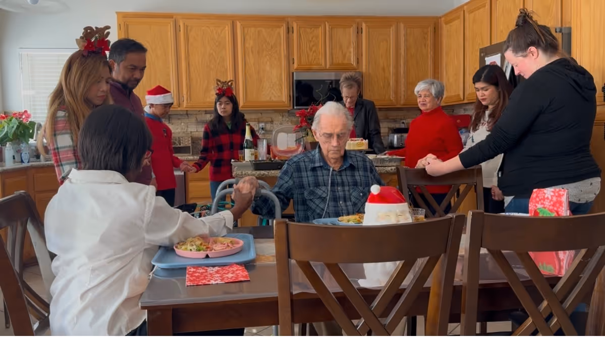 A group of people gathered in a kitchen around a dining table, holding hands in a circle. The setting appears festive with some individuals wearing Christmas-themed accessories like Santa hats and reindeer antlers. An elderly man is seated at the table with a meal in front of him, while others stand around him in a supportive gesture.