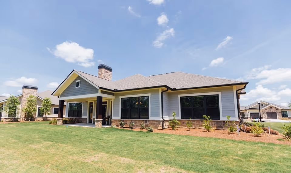 Exterior view of a single-story residential building with gray siding, stone accents, large windows, and a covered porch. The building is surrounded by a well-maintained lawn and small shrubs under a partly cloudy blue sky.