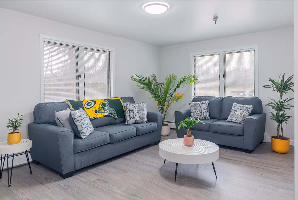 A bright living room with two gray sofas adorned with patterned pillows and a Green Bay Packers blanket. There are three potted plants placed around the room, including one on a small round side table and another on a white round coffee table with black legs. The room has light-colored wood flooring and two large windows with blinds, allowing natural light to enter.