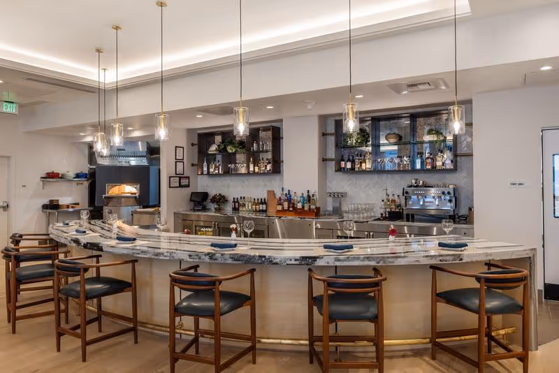 A modern curved bar area with a marble countertop and six wooden bar stools with black cushions. Behind the bar are shelves stocked with various bottles of alcohol, glassware, and a coffee machine. The space is well-lit with pendant lights hanging from the ceiling and has a clean, contemporary design.