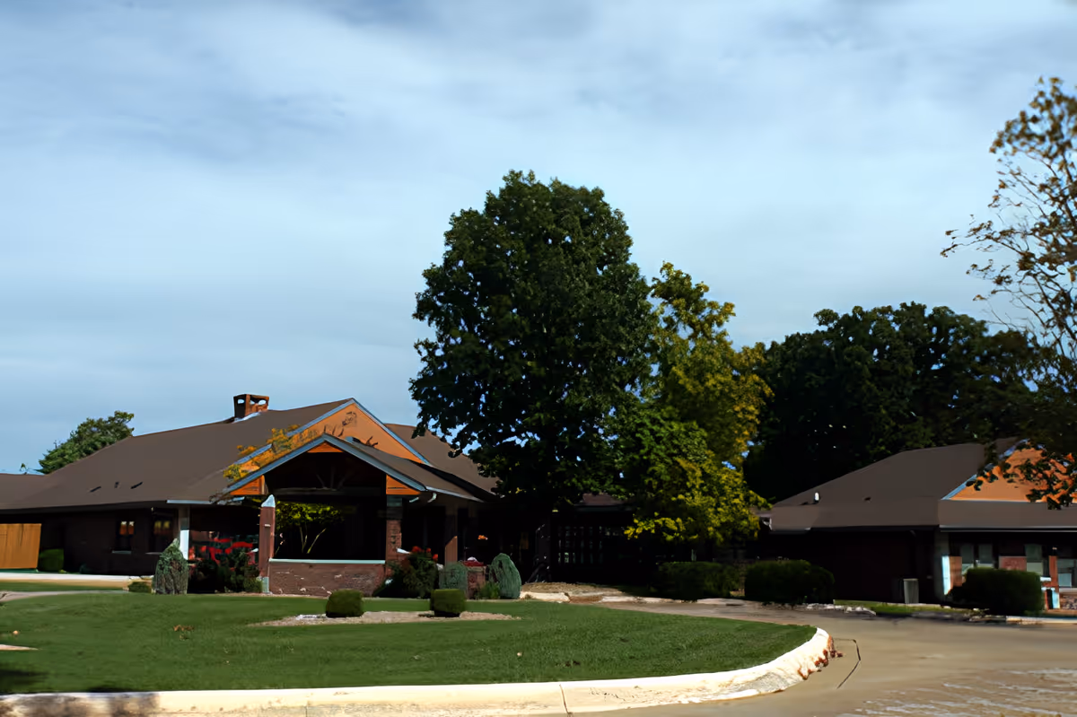 Exterior view of a single-story brick building with a brown roof surrounded by green trees and a well-maintained lawn under a partly cloudy sky.