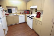 A compact kitchen with beige cabinets and drawers, a white refrigerator, dishwasher, and stove with four burners. There is a red microwave and a red coffee maker on the countertops. The floor is wooden, and the walls are light-colored.
