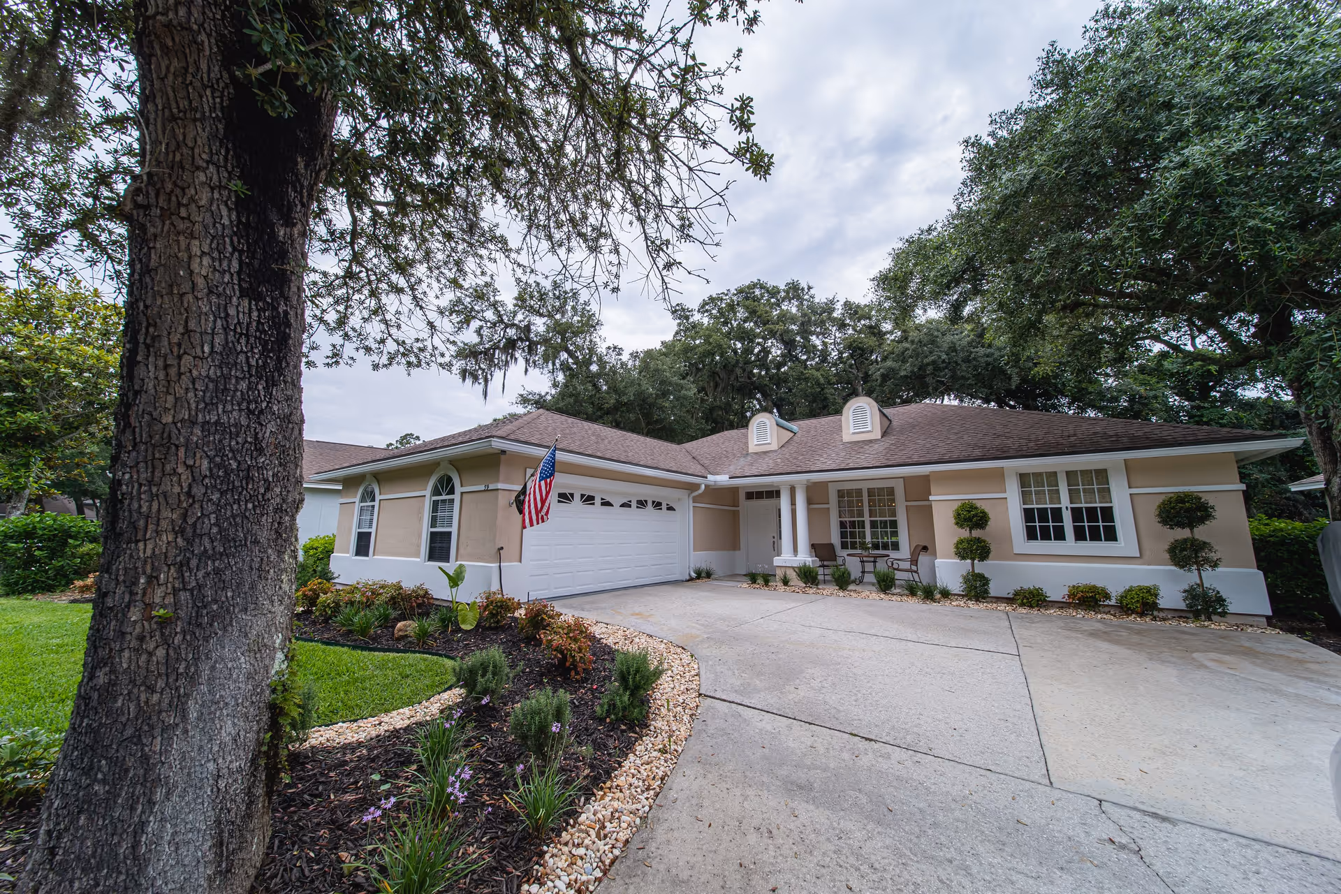 Exterior view of a single-story residential building with a beige facade and white trim, featuring a two-car garage with an American flag mounted beside it. The driveway is concrete and curves slightly, bordered by landscaped garden beds with small shrubs, flowers, and mulch. Large trees surround the property under a cloudy sky.
