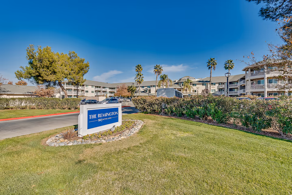 Exterior view of The Remington Senior Living facility with a clear blue sky, green lawn, palm trees, and a sign displaying the facility's name near the entrance driveway.