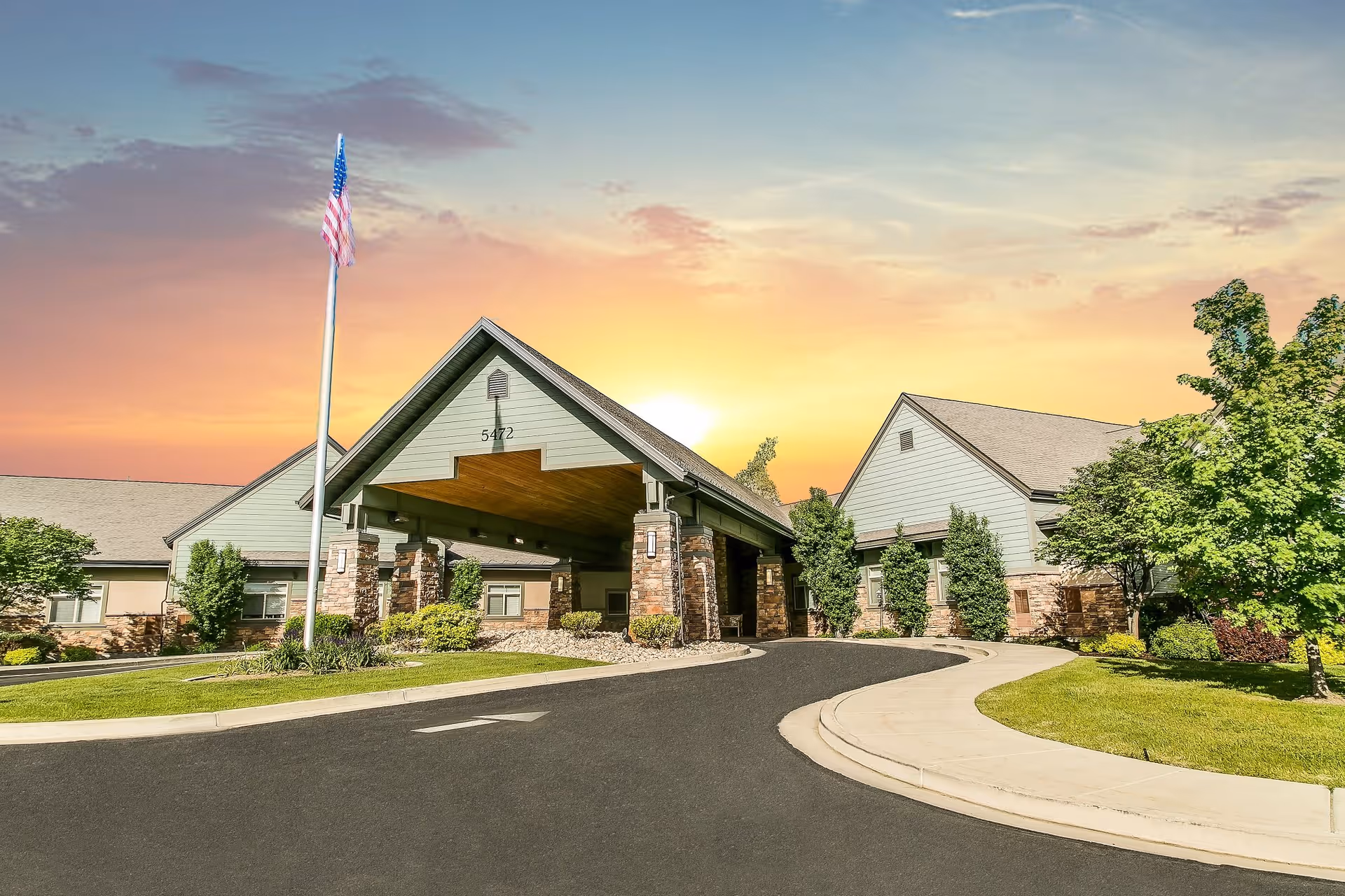 Exterior view of Legacy Village of Taylorsville building at sunset with a curved driveway, an American flag on a flagpole, and surrounding greenery including trees and shrubs.
