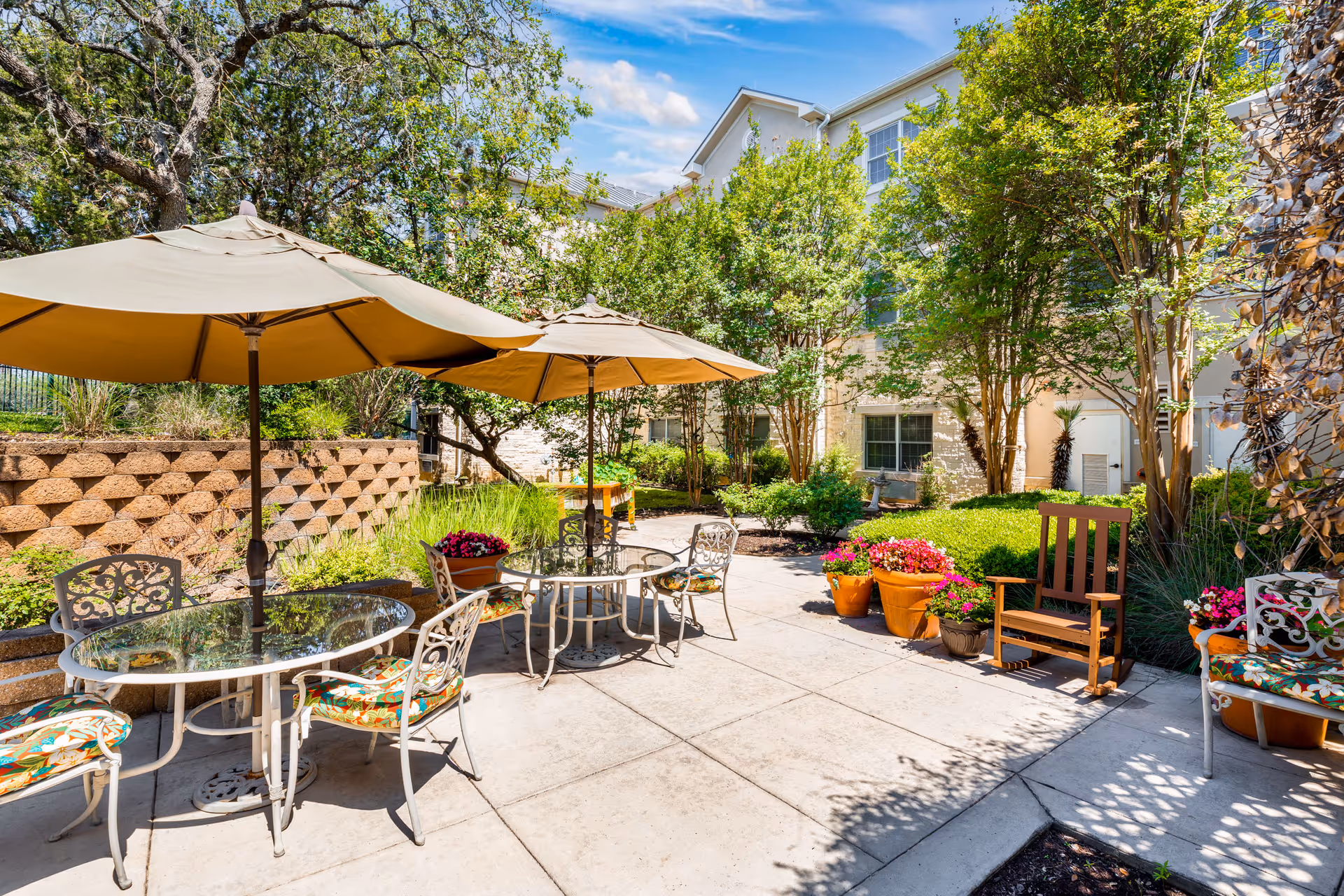Outdoor patio area at The Waterford at Shavano Park with two round glass tables, each shaded by large beige umbrellas. The tables are surrounded by metal chairs with floral cushions. The patio is surrounded by lush greenery, potted flowers, and trees, with a building visible in the background under a blue sky with some clouds.