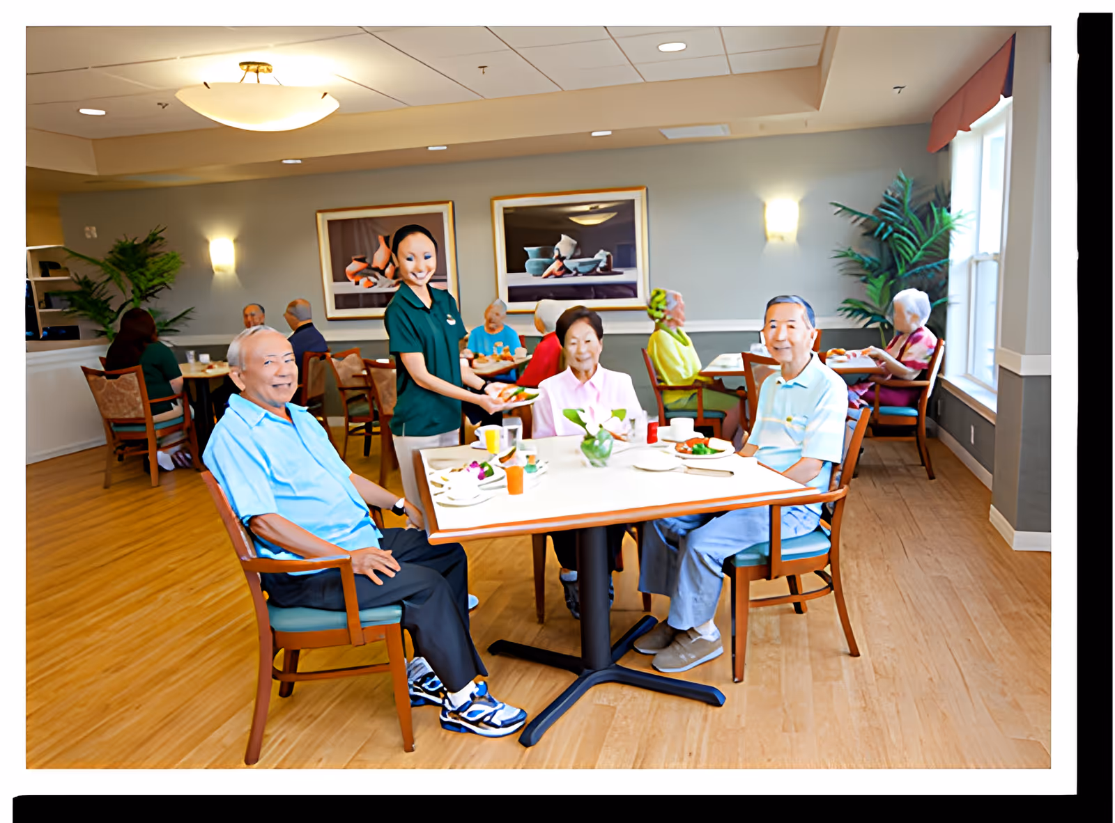 A dining room in a senior living facility with elderly residents seated at tables enjoying their meals. A staff member is serving food to a table where three elderly individuals are seated. The room has wooden flooring, framed artwork on the walls, and large windows letting in natural light.