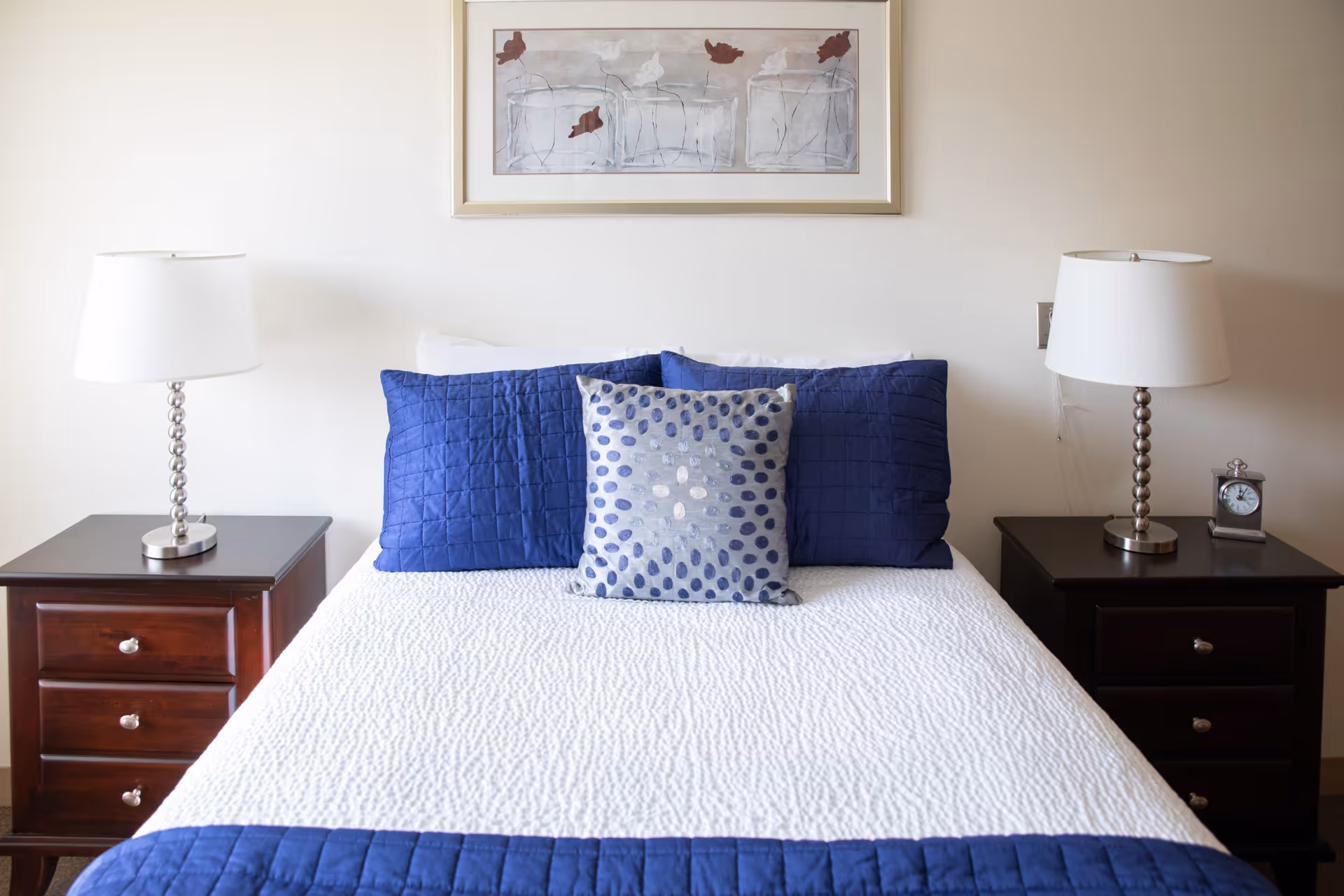 A neatly made bed with a white quilt and blue pillows, flanked by two dark wooden nightstands each with a silver lamp. A framed artwork featuring birds hangs on the wall above the bed.