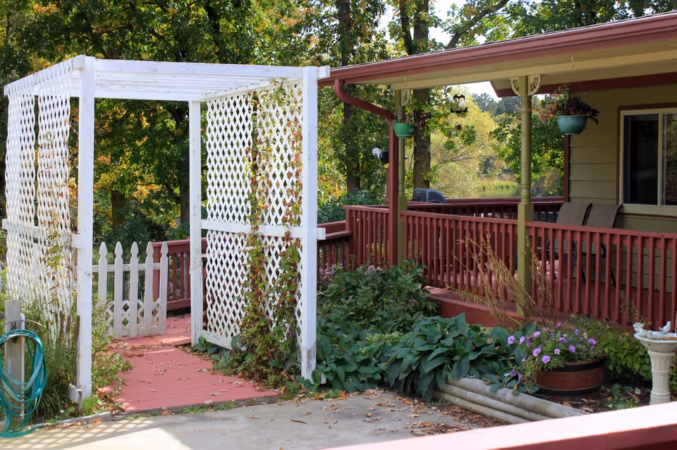Outdoor garden area with a white lattice pergola covered in climbing plants, a red wooden railing, green shrubs, potted flowers, and a house porch with hanging planters.