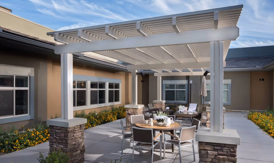 Outdoor courtyard with a white pergola over a patio seating area including a round table, chairs, and surrounding landscaping.