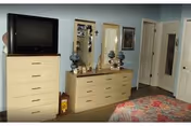 A bedroom with a dresser and a chest of drawers. On top of the chest of drawers is a television. The dresser has various decorative items including lamps and framed photos. There is a bed partially visible with a colorful quilt. The room has light-colored walls and doors.