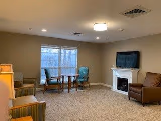 A cozy living room with beige walls and carpet, featuring a flat-screen TV mounted above a white fireplace. There are several chairs arranged around the room, including two green armchairs near a window with blinds, a brown leather armchair near the fireplace, and another chair partially visible on the left side. The room is softly lit by ceiling lights.