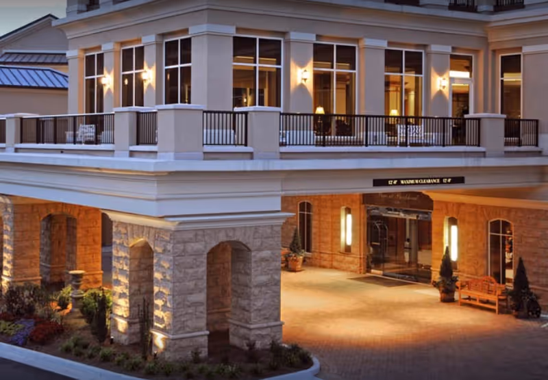 Exterior view of a senior living facility entrance at dusk with stone pillars, warm lighting, large windows, and a covered drop-off area. There are potted plants and a bench near the entrance.