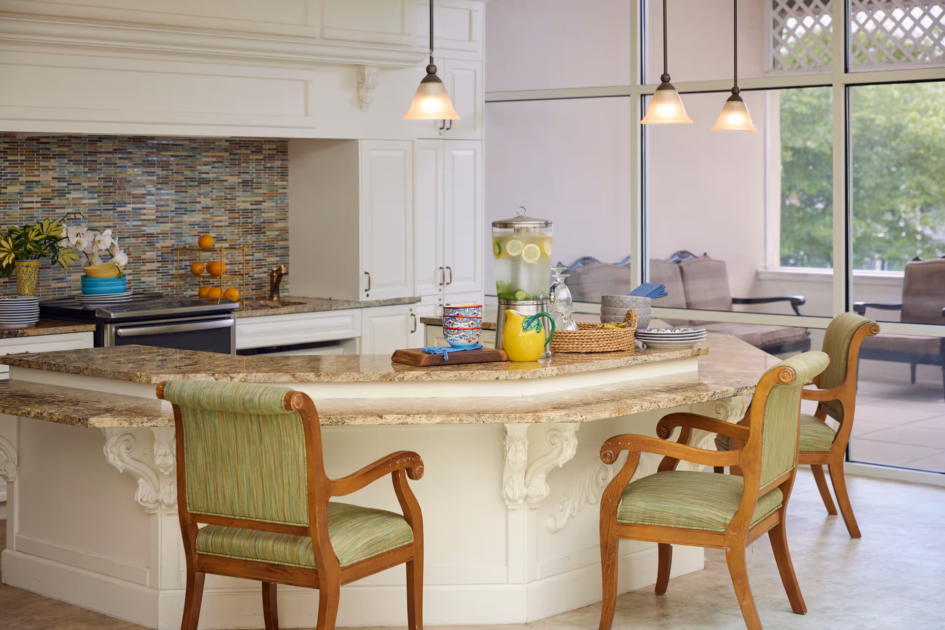 A bright kitchen area with a curved granite countertop island surrounded by three wooden chairs with green upholstered seats and backs. The kitchen features white cabinetry, a mosaic tile backsplash, and stainless steel appliances. On the countertop, there is a glass beverage dispenser with lemon and lime slices, colorful bowls, a yellow pitcher, and a basket with napkins. Large windows in the background reveal an outdoor seating area with cushioned chairs.