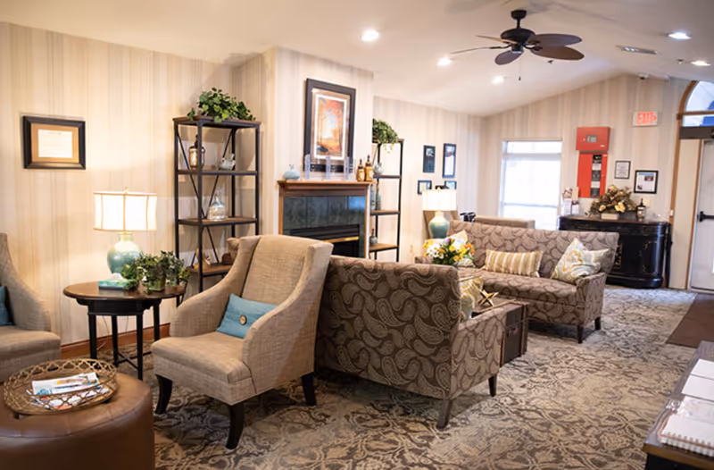 Cozy lounge area with upholstered chairs and sofas around a fireplace, side tables, lamps, and decorative shelving.