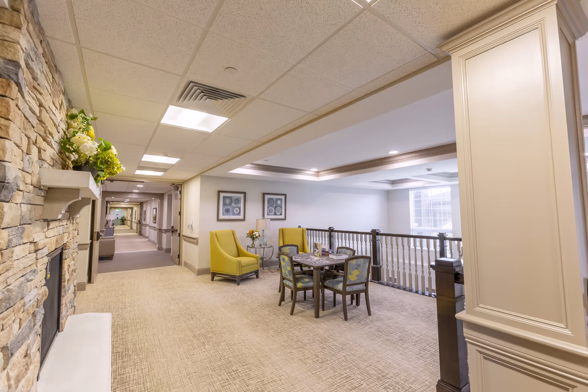 A bright and spacious common area in a senior living facility featuring a stone fireplace on the left, a seating area with two yellow armchairs and a small round table with flowers, and a square table with four chairs. The area has beige carpeting, framed artwork on the walls, and a railing overlooking a lower level with large windows letting in natural light.