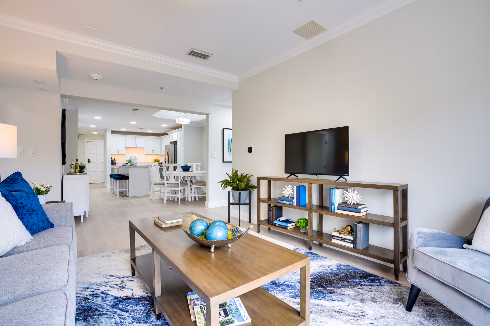 A modern living room with a gray sofa adorned with blue and white pillows, a wooden coffee table with decorative blue spheres, a patterned blue and white rug, a wooden TV stand with books and decor items, and a flat-screen TV mounted on the wall. In the background, a dining area with a white dining table and chairs is visible, along with a kitchen featuring white cabinetry and stainless steel appliances.
