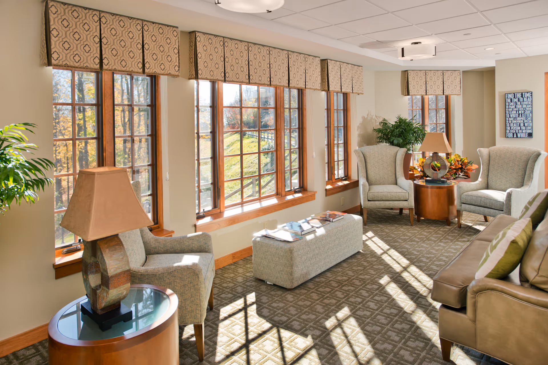 Sunlit sitting room with large multi-pane windows, upholstered chairs and sofa, side tables with lamps, and decorative plants.