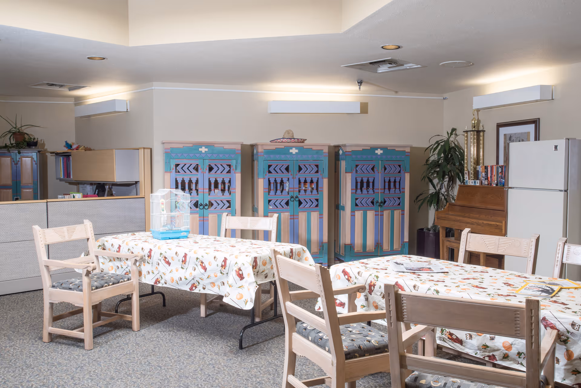 A common area in a senior living facility with two tables covered in patterned tablecloths, surrounded by wooden chairs with cushioned seats. On one table, there is a small birdcage. In the background, there are three colorful cabinets with geometric designs, a piano with a large trophy on top, a refrigerator, and some plants. The room has a carpeted floor and soft lighting.