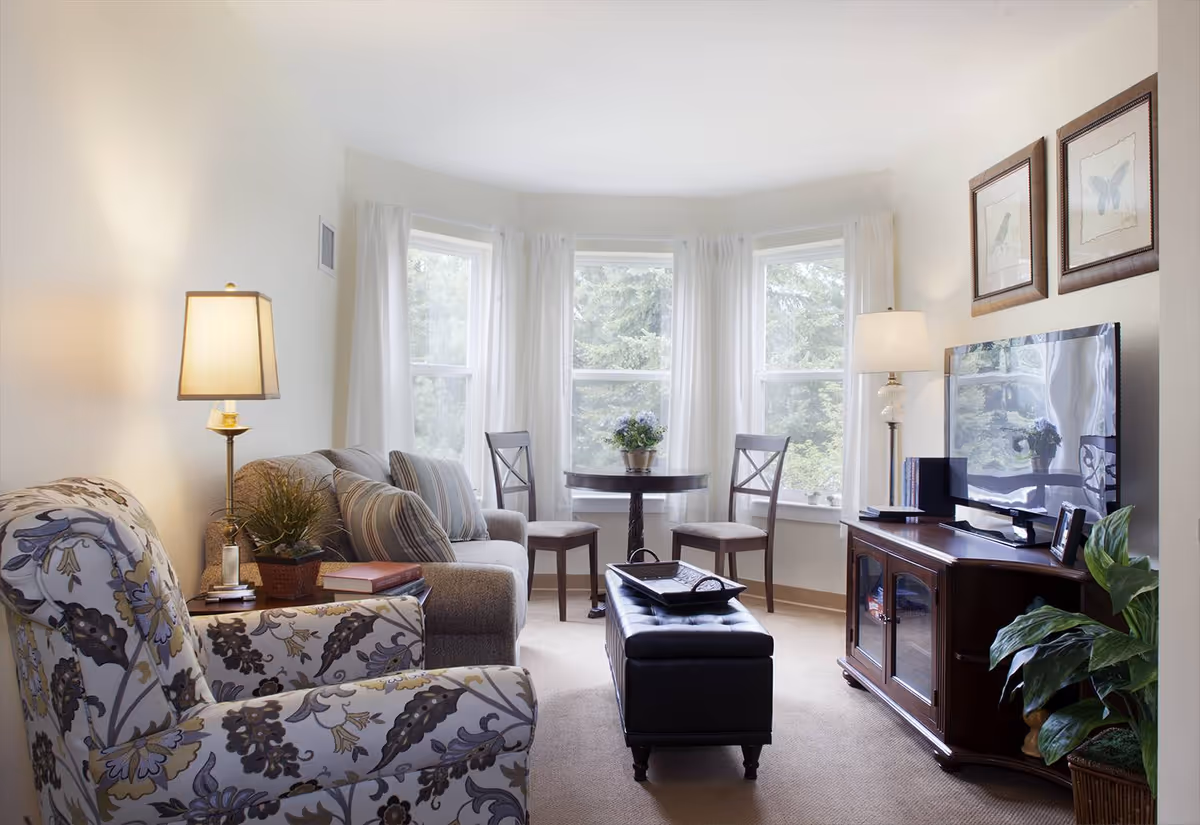 A bright and cozy living room with a floral-patterned armchair, a beige sofa with striped pillows, a dark wooden TV stand with a flat-screen TV, two framed pictures on the wall, a round table with two chairs in front of three large windows with white curtains, and two lamps providing warm lighting.