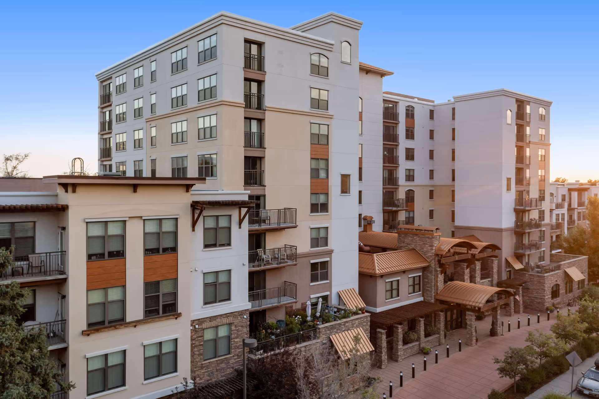 Exterior view of a multi-story senior living facility building with balconies, large windows, and a covered entrance area. The building features a combination of stone and stucco finishes with wood accents, surrounded by trees and a paved walkway.