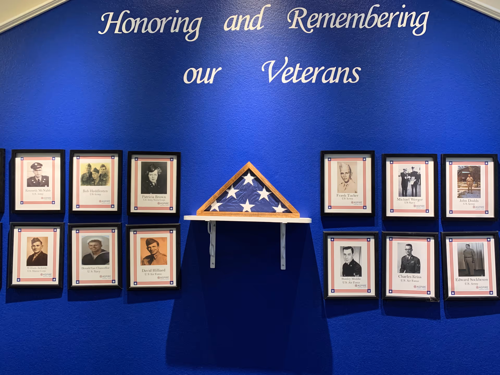 Memorial wall with framed veteran photos and a folded American flag on a shelf beneath the words "Honoring and Remembering our Veterans" on a blue wall.