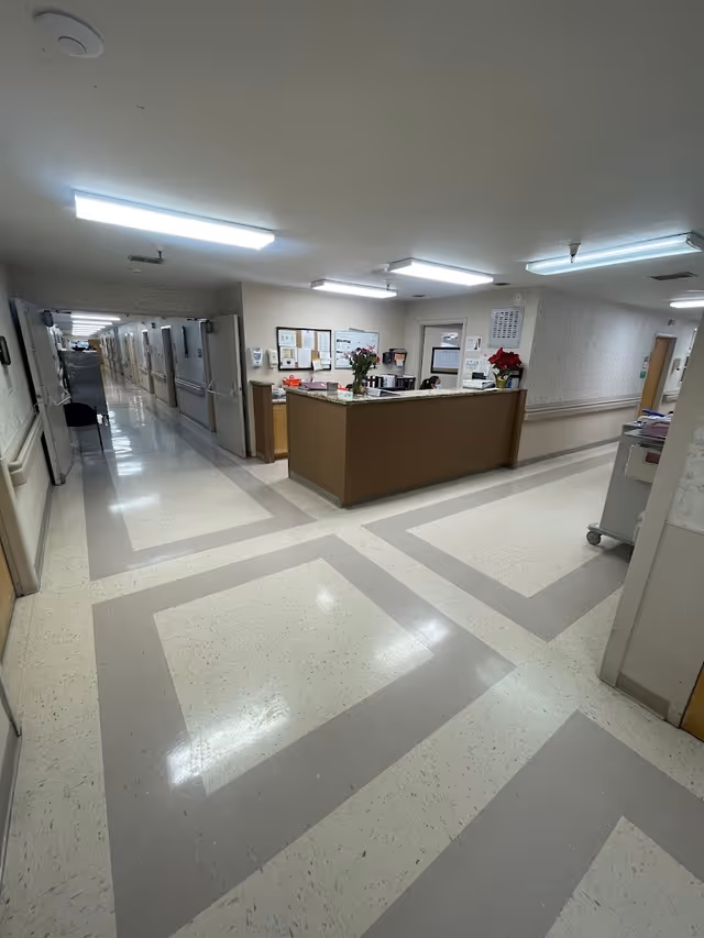 Interior view of a healthcare facility hallway with a nurse's station in the center. The floor has a patterned design with light and dark tiles. The hallway extends to the left with doors along the walls, and the nurse's station has bulletin boards, flowers, and office supplies on the counter.