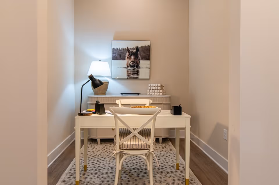 A small, cozy office nook with a white desk and chair on a patterned rug. On the desk are a black desk lamp, a pencil holder, and some small items. Behind the desk is a sideboard with a table lamp and decorative objects. A framed picture of a horse partially submerged in water hangs on the wall above the sideboard.