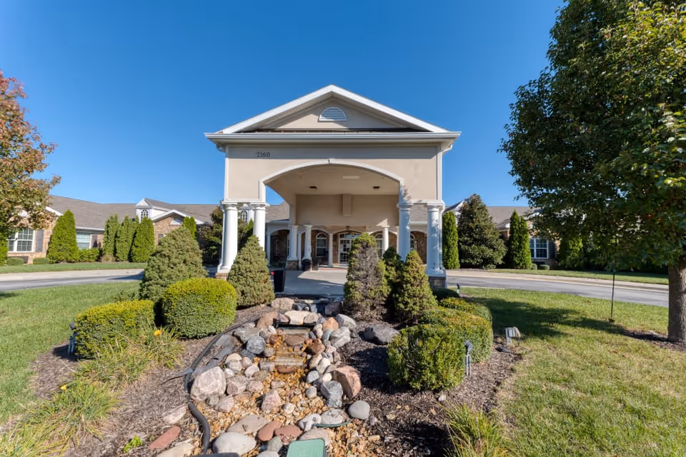 Front entrance of a senior living facility with a covered porte-cochère, white columns, landscaped rock water feature and surrounding shrubs under a clear blue sky.