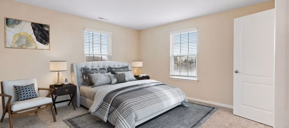 A bright bedroom with beige walls and carpeted floor featuring a large bed with gray and white bedding, two bedside tables with lamps, a wooden armchair with a patterned cushion, two windows with white blinds, and a modern abstract painting on the wall.