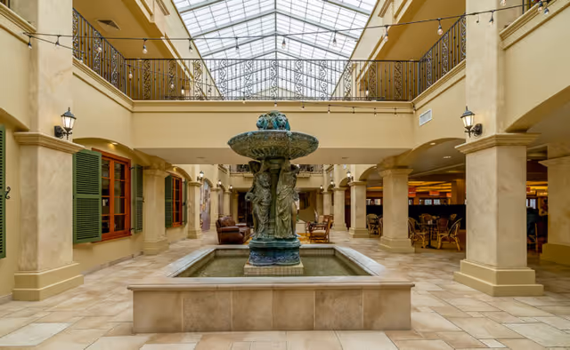 Indoor courtyard area with a large decorative fountain in the center featuring sculpted figures. The space has beige tiled floors, cream-colored columns, and a glass ceiling allowing natural light to fill the area. There are seating areas with chairs and tables along the sides, and green window shutters on the left wall.
