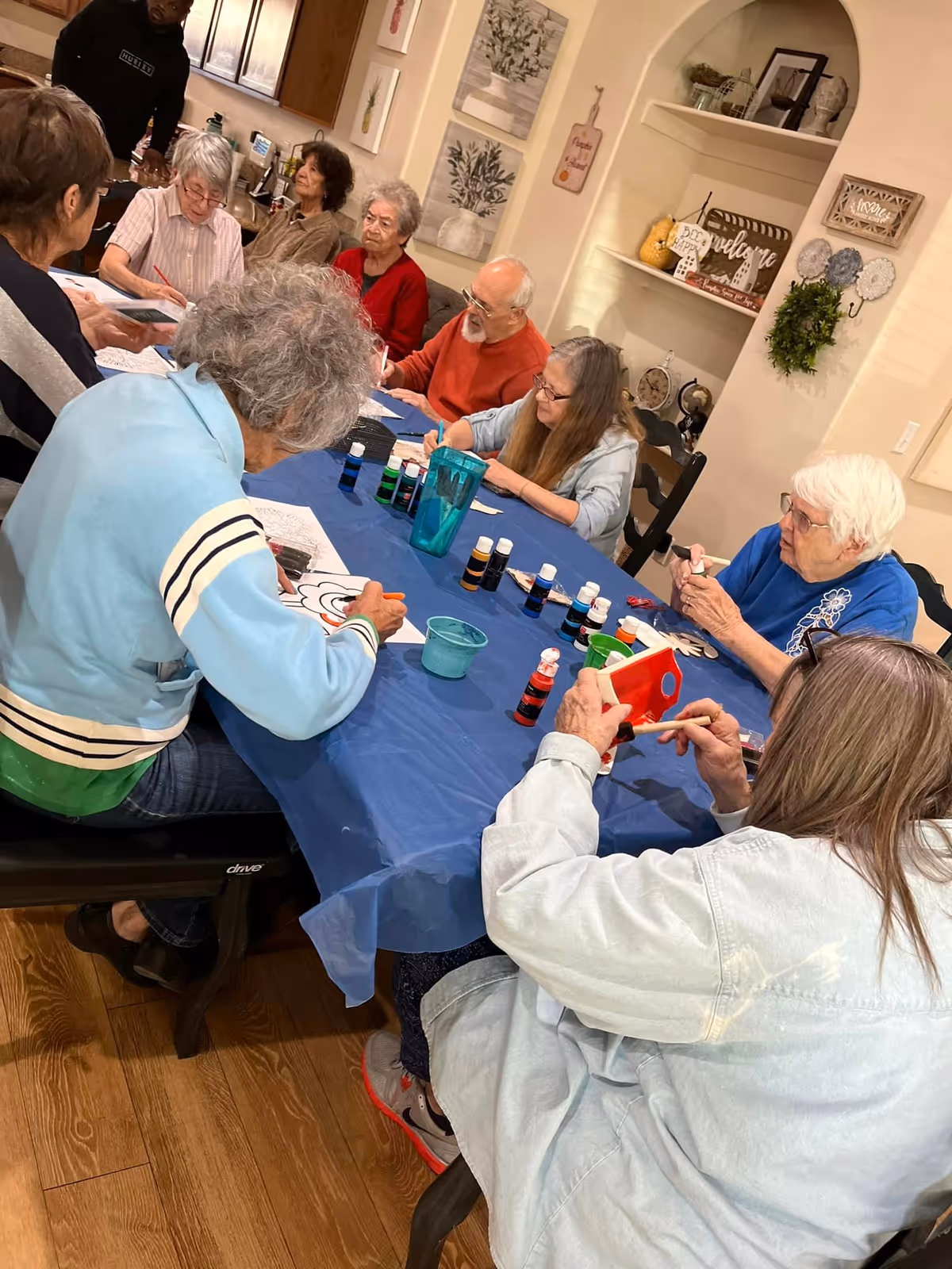 A group of elderly people sitting around a table covered with a blue tablecloth, engaging in arts and crafts activities such as painting and coloring. The room has wooden floors, decorated walls with framed pictures and shelves holding decorative items.