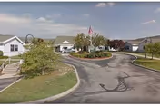 Exterior view of a single-story health and rehabilitation facility with a circular driveway, landscaped greenery, and an American flag in front of the entrance under a partly cloudy sky.