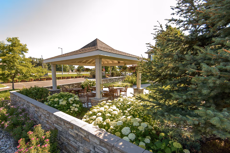 Outdoor gazebo with a shingled roof surrounded by a stone wall and lush greenery including white hydrangeas and pine trees. Several wooden tables and chairs are arranged under the gazebo, with a clear blue sky in the background.