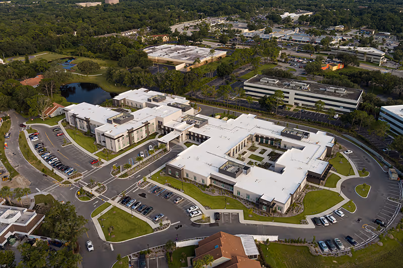 Aerial view of a U-shaped senior living facility with white roofs, inner courtyards, parking lots and surrounding greenery.