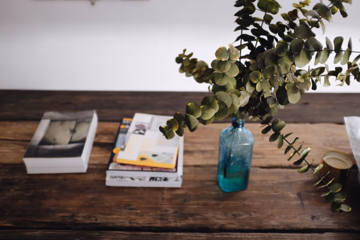 A rustic wooden table with a blue glass bottle holding green eucalyptus branches, a stack of books and magazines, and a small cup on the right side.