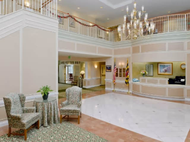 Spacious and well-lit senior living facility lobby with high ceilings, a chandelier, two upholstered chairs with a small table and plant, a reception desk, and flags displayed near the entrance.