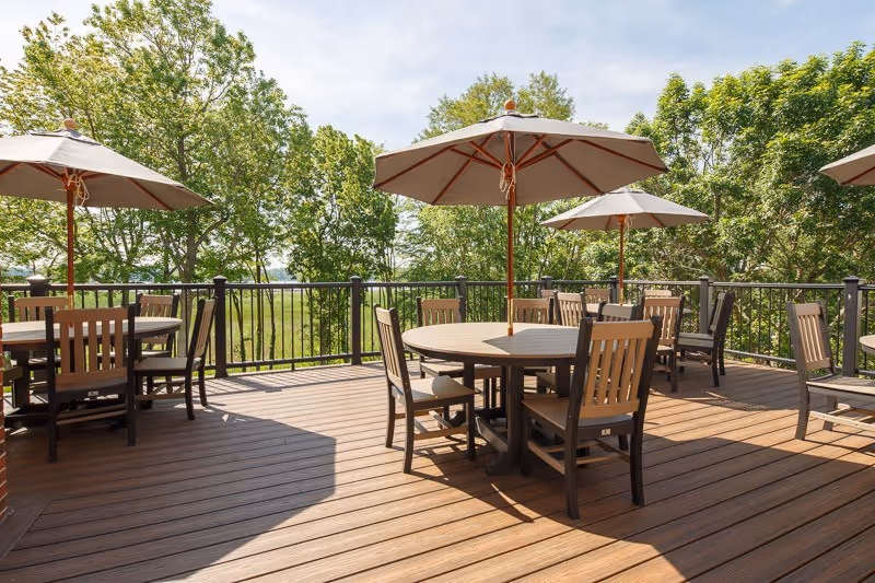 Outdoor wooden deck area with round tables and chairs, each table shaded by a large umbrella. The deck is surrounded by a black metal railing and lush green trees are visible in the background under a partly cloudy sky.