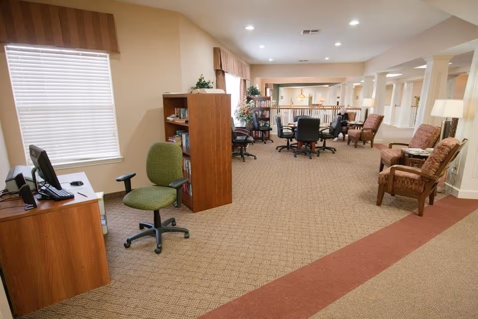 Interior view of a senior living facility common area with carpeted floor, several armchairs, a round table with office chairs, bookshelves, a desk with a computer, and large windows with blinds and valances.