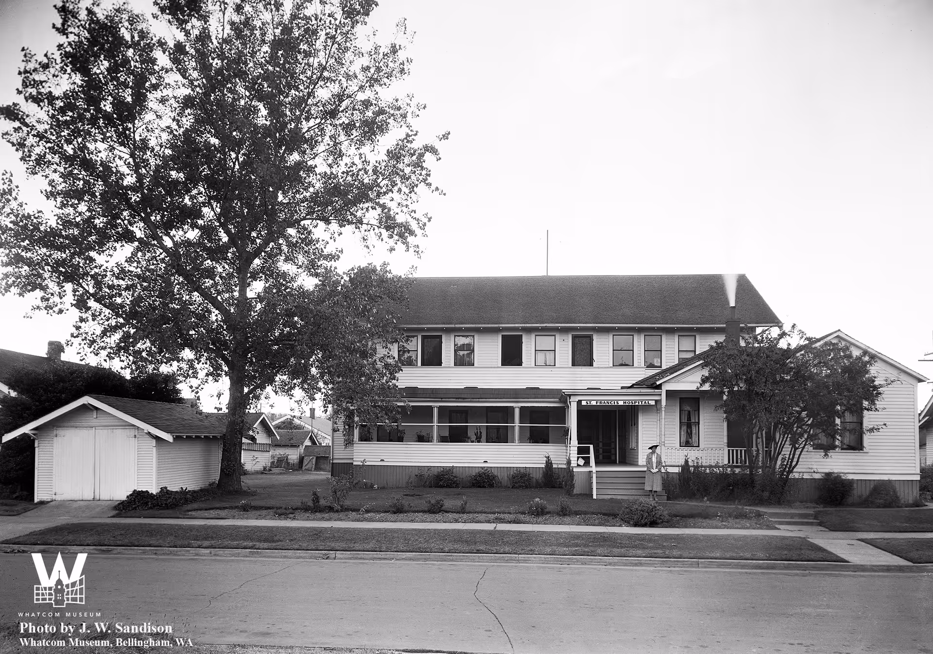 Black-and-white photograph of a two-story house-like building with a front porch, a large tree, and a detached garage facing a street.