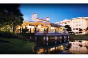 Well-lit multi-story senior living building with a covered porch reflected in a pond at twilight.