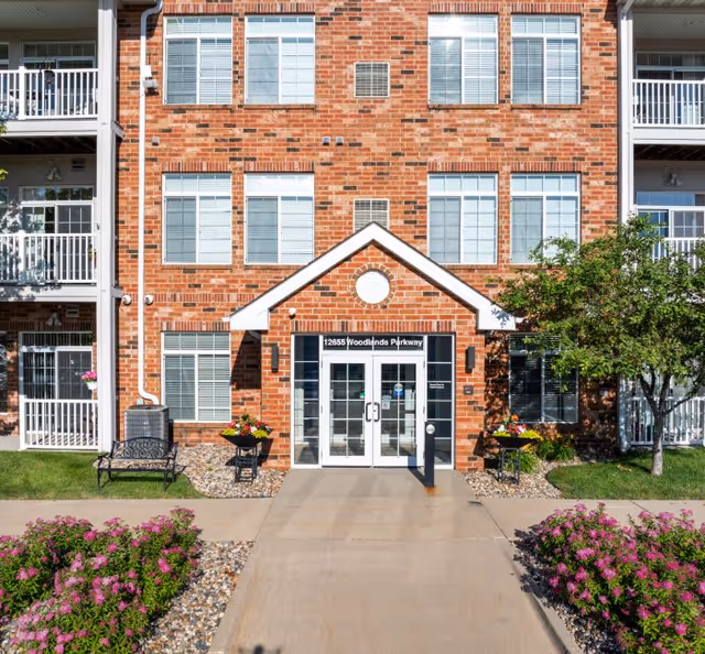Brick senior living building with glass doors, landscaping, and benches near the entrance.