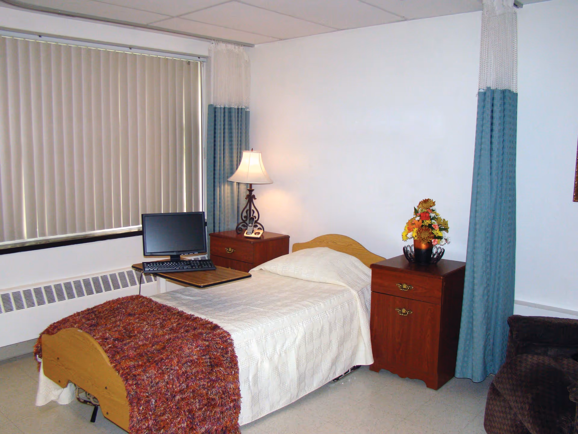 A simple nursing center bedroom with a single bed covered in a white bedspread and a reddish-brown throw blanket. There are two wooden nightstands on either side of the bed, one with a lamp and the other with a flower arrangement. A computer monitor and keyboard are placed on a small table attached to the bed. The room has vertical blinds covering the window and blue privacy curtains hanging from the ceiling.