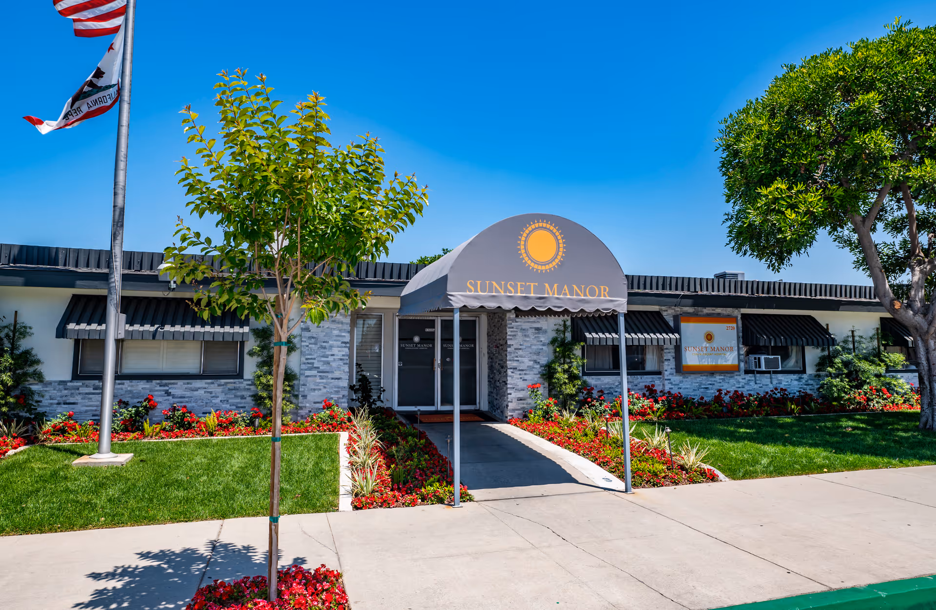 Exterior front view of Sunset Manor facility with a covered entrance, well-maintained landscaping including green grass, red flowers, and trees, and an American flag on a flagpole against a clear blue sky.