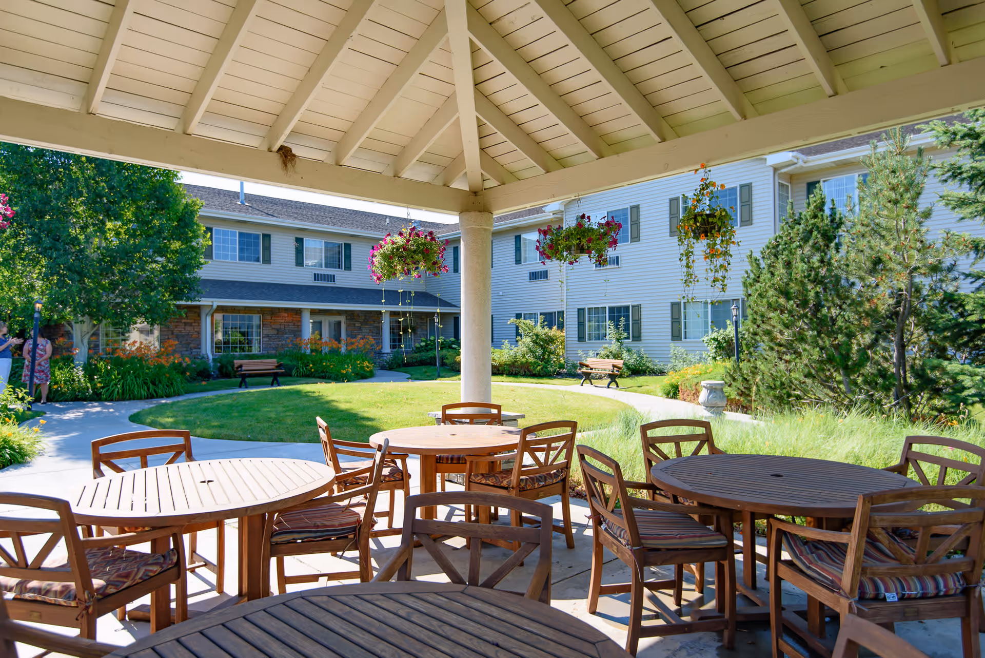 Outdoor covered seating area with wooden tables and chairs, hanging flower baskets, and a view of a garden with green grass, trees, and a multi-story building in the background.
