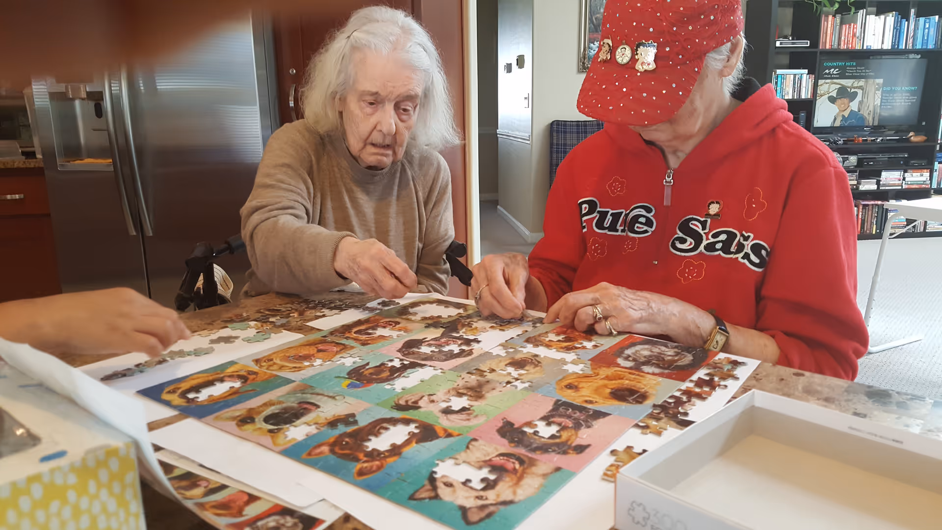 Two elderly women sitting at a table working together on assembling a colorful jigsaw puzzle featuring various dog images. One woman is wearing a red jacket and red cap, while the other is in a brown sweater. The setting appears to be a cozy living room with a bookshelf and a television in the background.