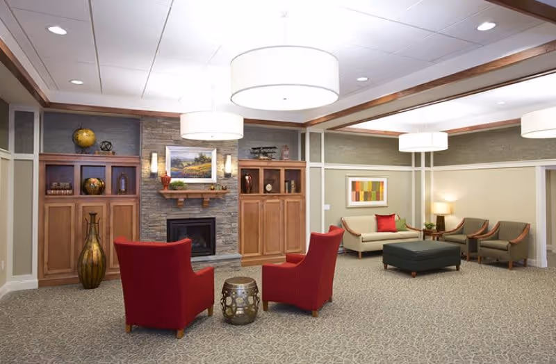 A spacious and well-lit living room area in an assisted living facility featuring two red armchairs facing a small decorative table, a stone fireplace with wooden cabinets on either side, a beige sofa with colorful cushions, two green armchairs, a large ottoman, and modern ceiling lights.