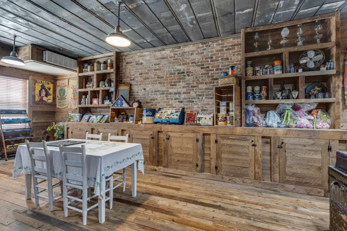 Rustic communal dining room with a white table and chairs in front of long wooden cabinets and shelves filled with jars and decor against an exposed brick wall.